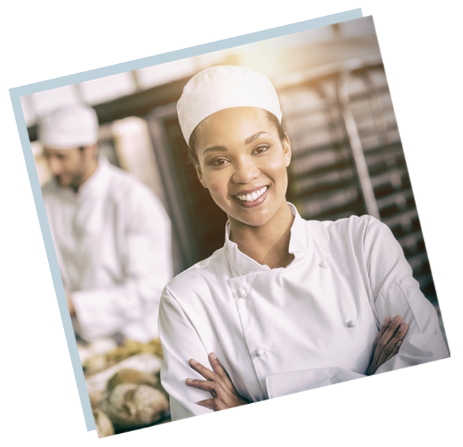 A commercial baker smiles with bread and coworkers in the background